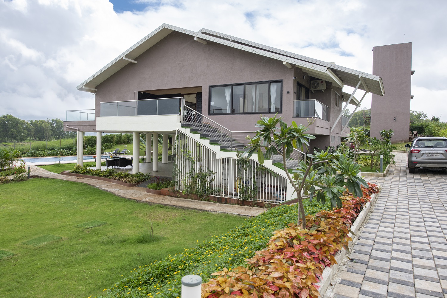 A modern luxury villa surrounded by a beautifully landscaped garden, featuring a spacious balcony, large glass windows, a private swimming pool, and a well-designed outdoor seating area, set in a peaceful farmhouse-style environment with a paved driv