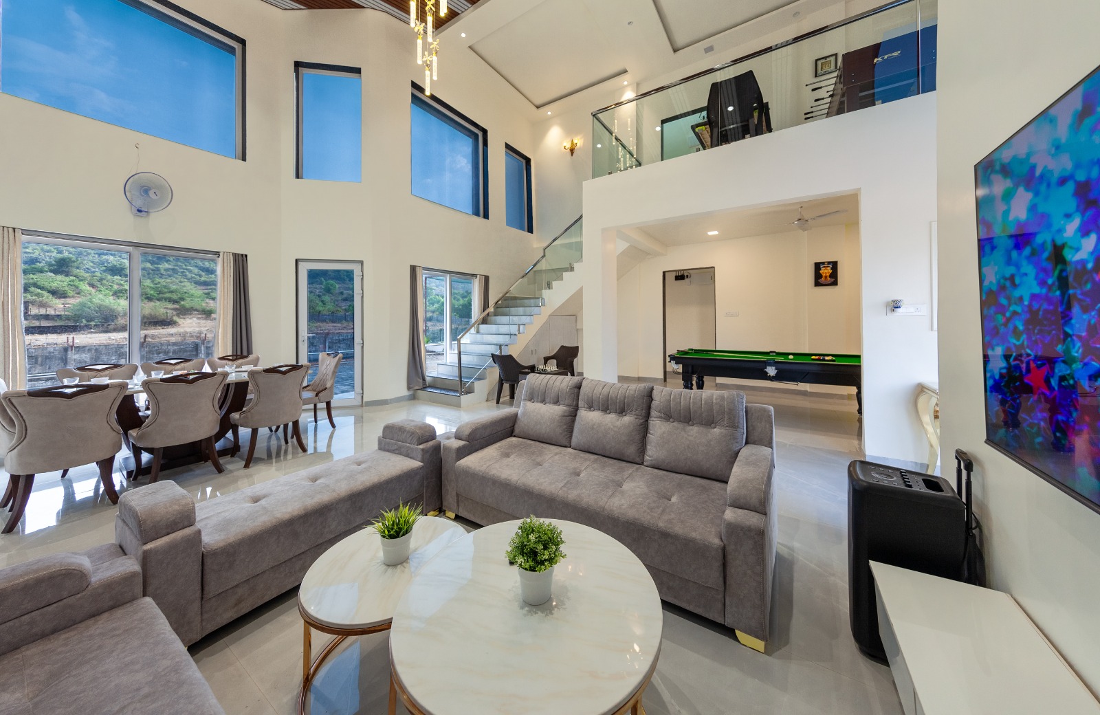 Modern luxury villa interior featuring a spacious double-height living room with grey sofas, a dining area, a glass staircase, and an indoor pool table designed with contemporary finishes and ample natural light.