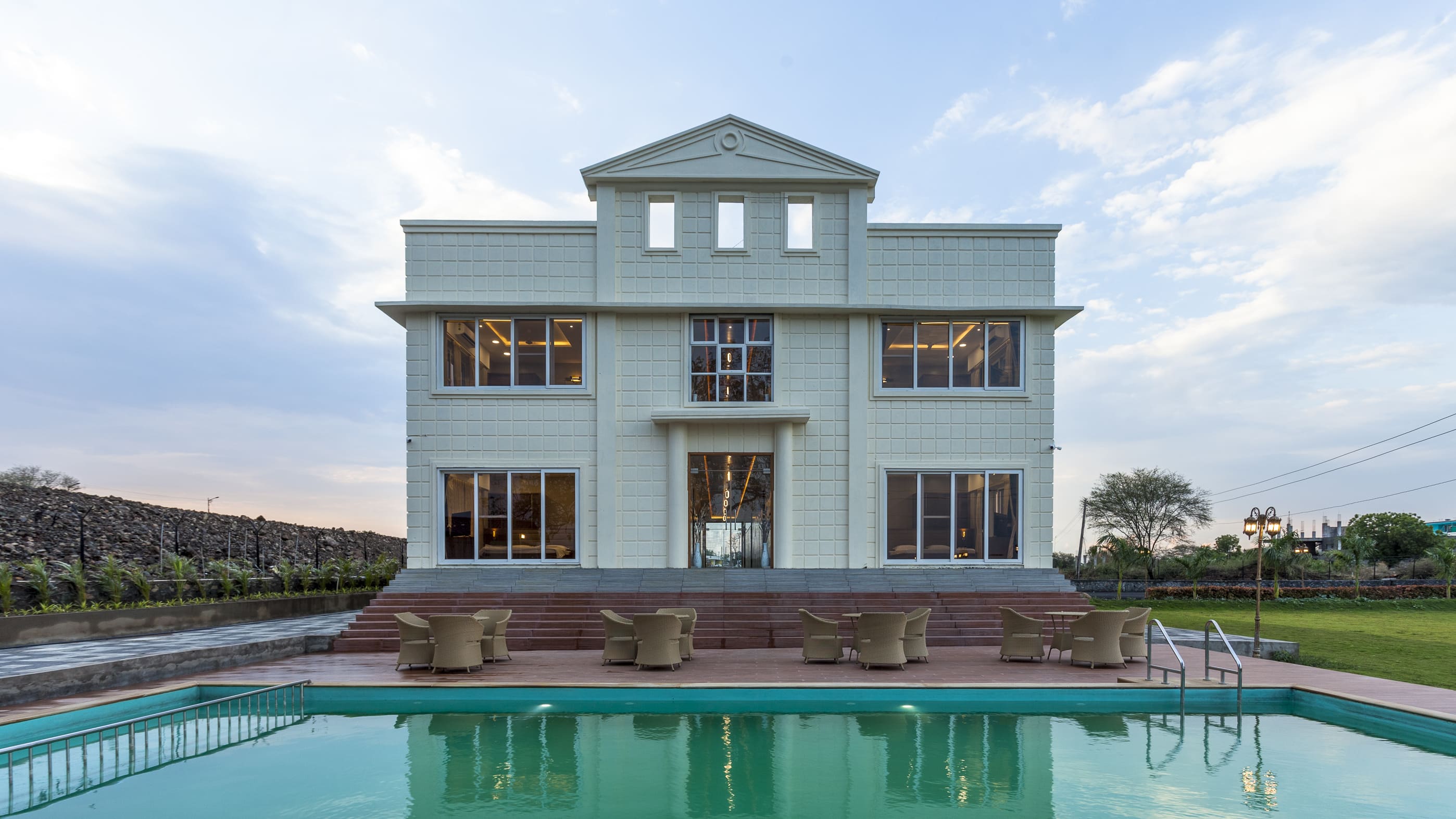A luxurious modern villa with a grand symmetrical façade, large glass windows, and elegant outdoor seating area overlooking a private swimming pool, set in a spacious landscaped property under a clear sky.